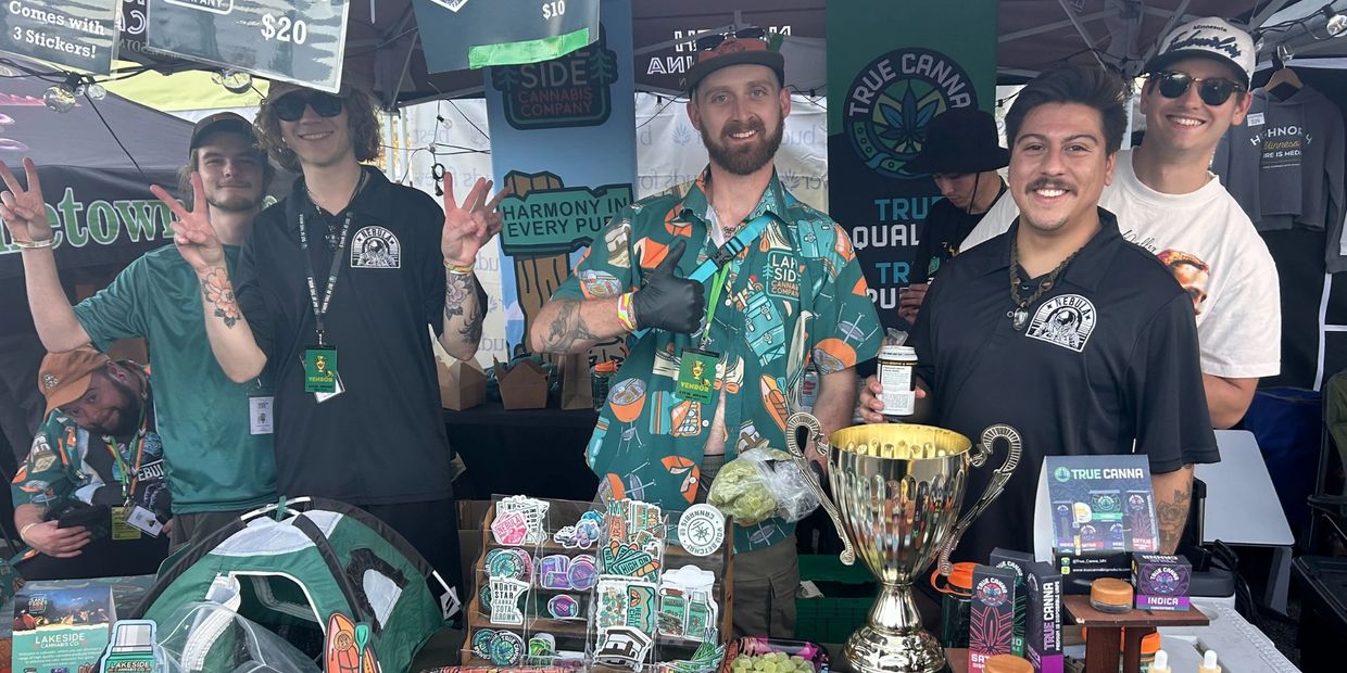 Group of smiling vendors posing at a colorful cannabis product booth with a large trophy.