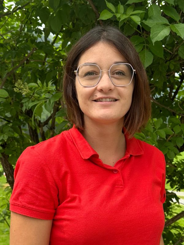 Woman in red polo shirt smiling outdoors with green leafy background.