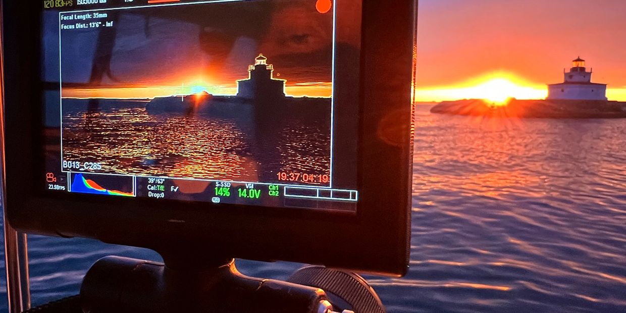 Sunset behind a lighthouse captured on a camera monitor by the water.