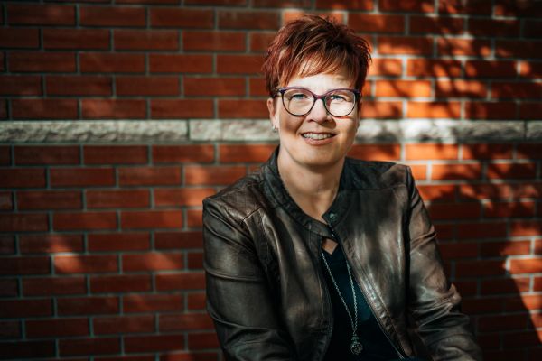 Smiling woman in glasses and leather jacket against brick wall.