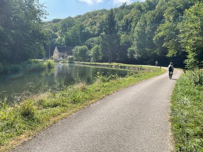 A cyclist rides on a paved path beside a calm river and lush greenery.