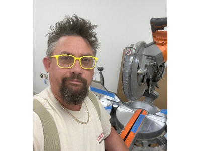 Man with yellow glasses posing near a miter saw in a workshop.