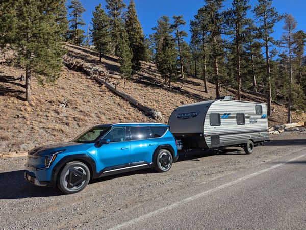 Blue electric SUV towing a travel trailer on a sunny forest road.
