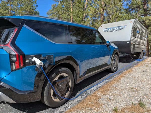 Blue electric SUV charging while towing a camper in a forested area.