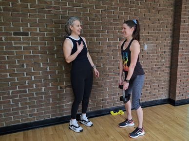 Two women in workout clothes chatting in a gym with dumbbells.