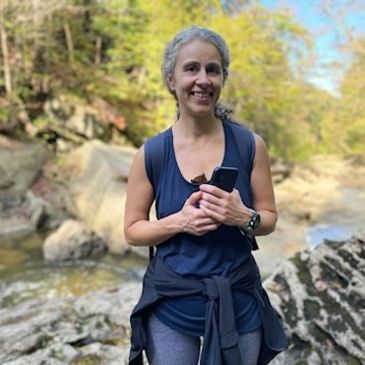 Smiling woman holding a phone outdoors near a rocky stream.