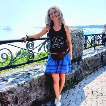 A woman smiling and posing by a stone wall with a scenic waterfront view.