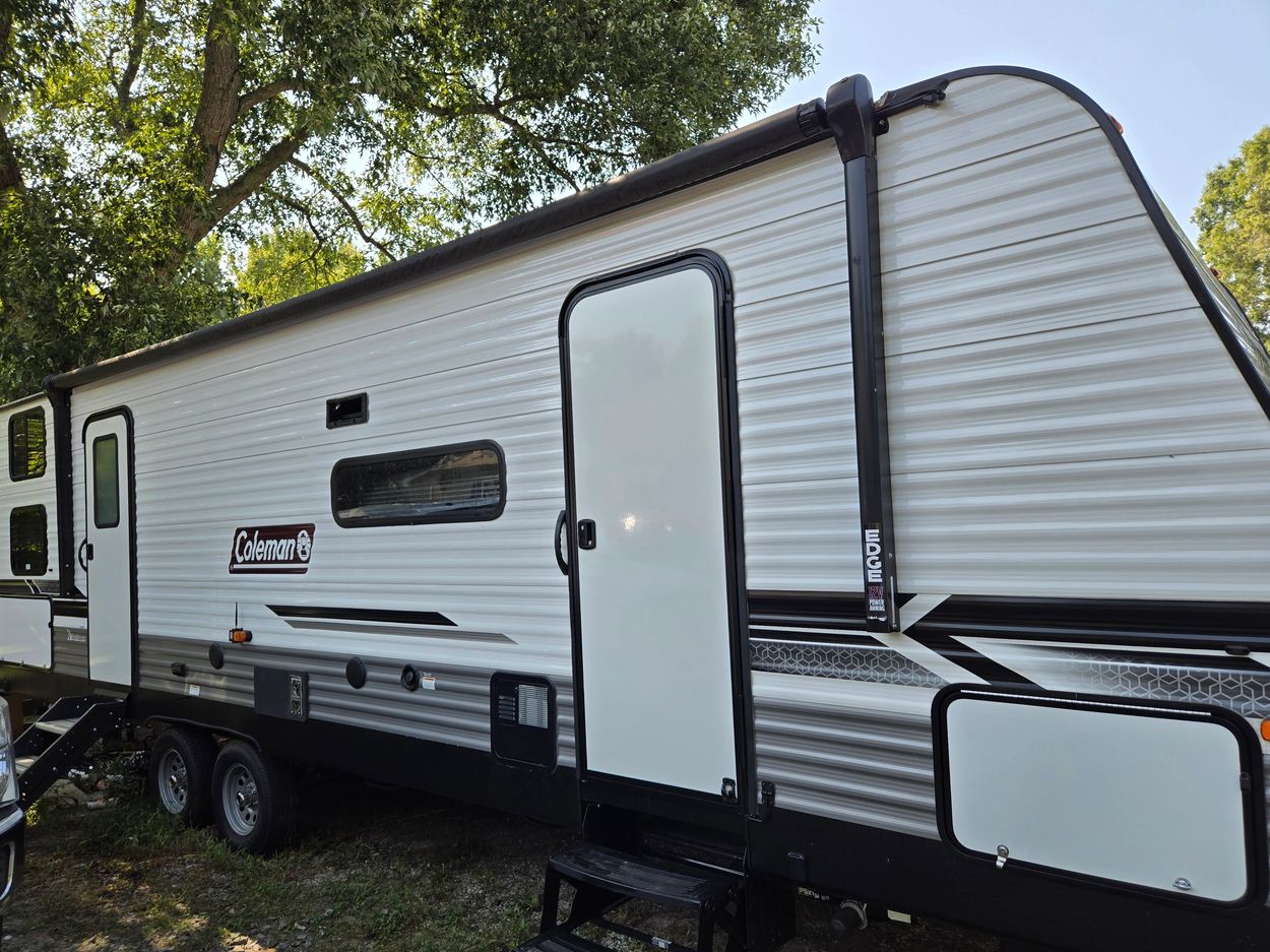 White Coleman travel trailer parked under a tree on grass.