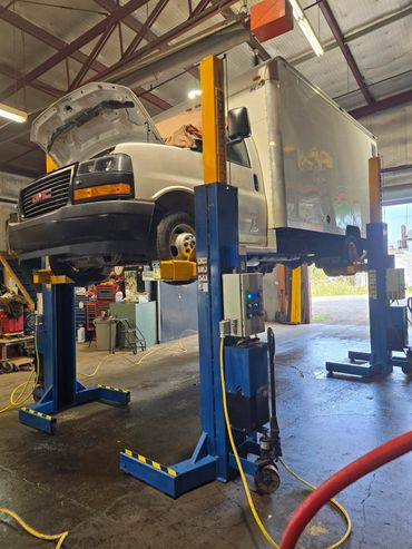A white GMC box truck lifted on a blue hydraulic lift in a garage.