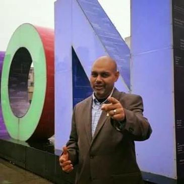 Man in a suit pointing in front of colorful large letters outdoors.
