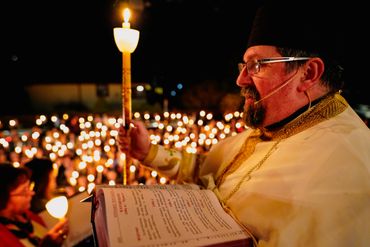 A priest holds a lit candle during a nighttime religious ceremony with many candles in the background.