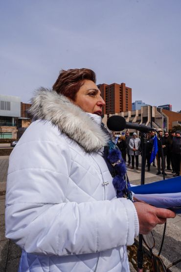 Woman speaking at outdoor event in a white coat.