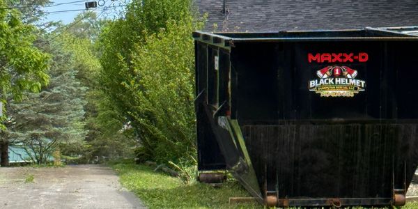 A large black dumpster labeled 'MAXX-D' and 'Black Helmet' sits on a driveway near green trees.