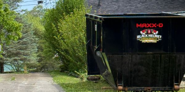 A large black dumpster labeled 'MAXX-D' and 'Black Helmet' sits on a driveway near green trees.