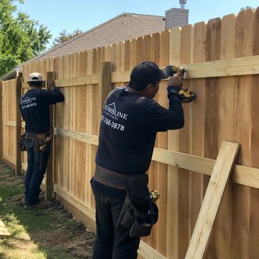 Two workers installing a wooden fence outdoors on a sunny day.
