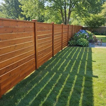 A wooden fence casting shadows on a green lawn beside a garden with chairs.