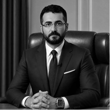 A serious man in a suit sitting at a desk with hands clasped.