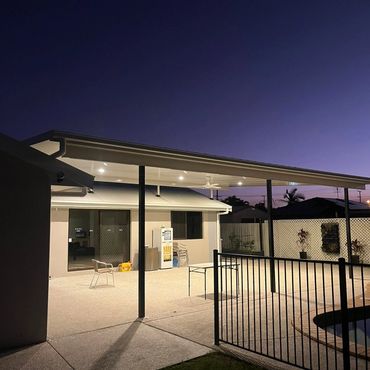 Evening view of a well-lit backyard patio with chairs and a table under a covered roof.