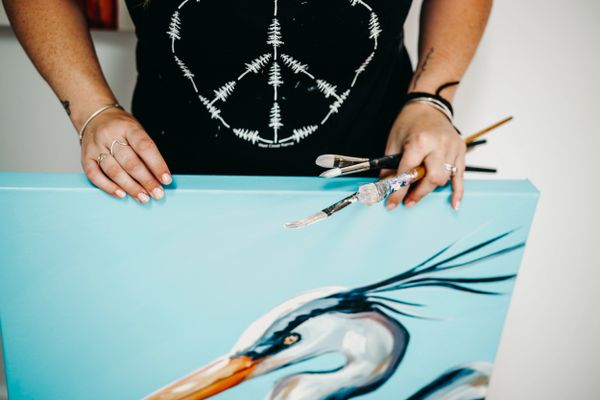 Artist holding paintbrushes and a painting of a bird on a blue canvas.