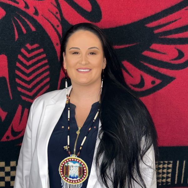 Smiling woman with long black hair wearing traditional beadwork necklace and white jacket.