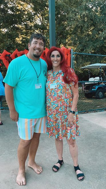 A smiling couple poses outdoors by a fence, dressed in colorful summer attire.