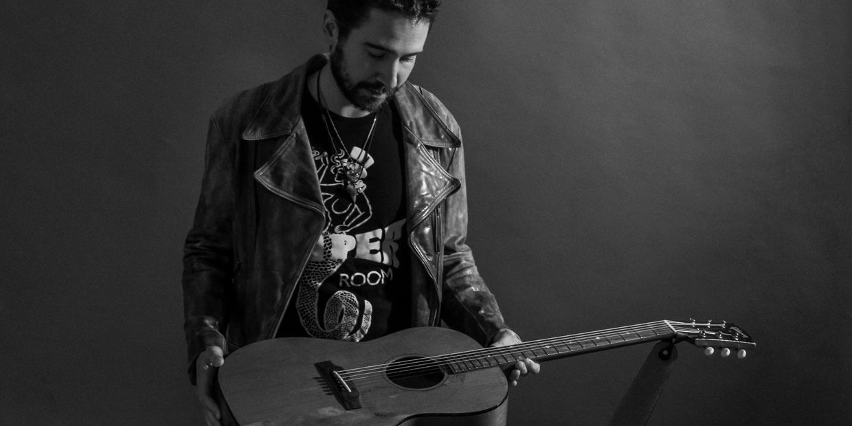 Man in leather jacket holding an acoustic guitar, looking down.