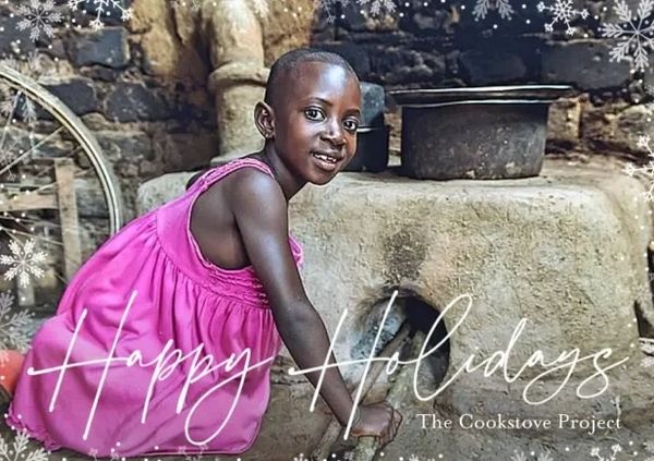 A smiling girl in a pink dress near a traditional cookstove.