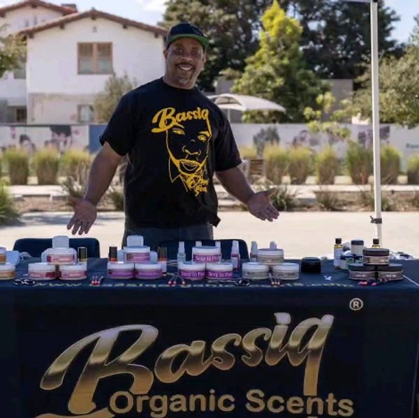 Man smiling behind a table of Bassiq Organic Scents products at an outdoor market.