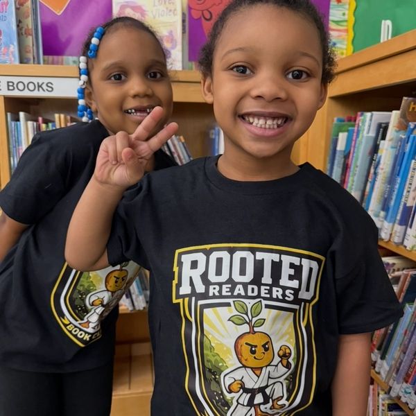 Two smiling children in matching 'Rooted Readers' shirts at a library.