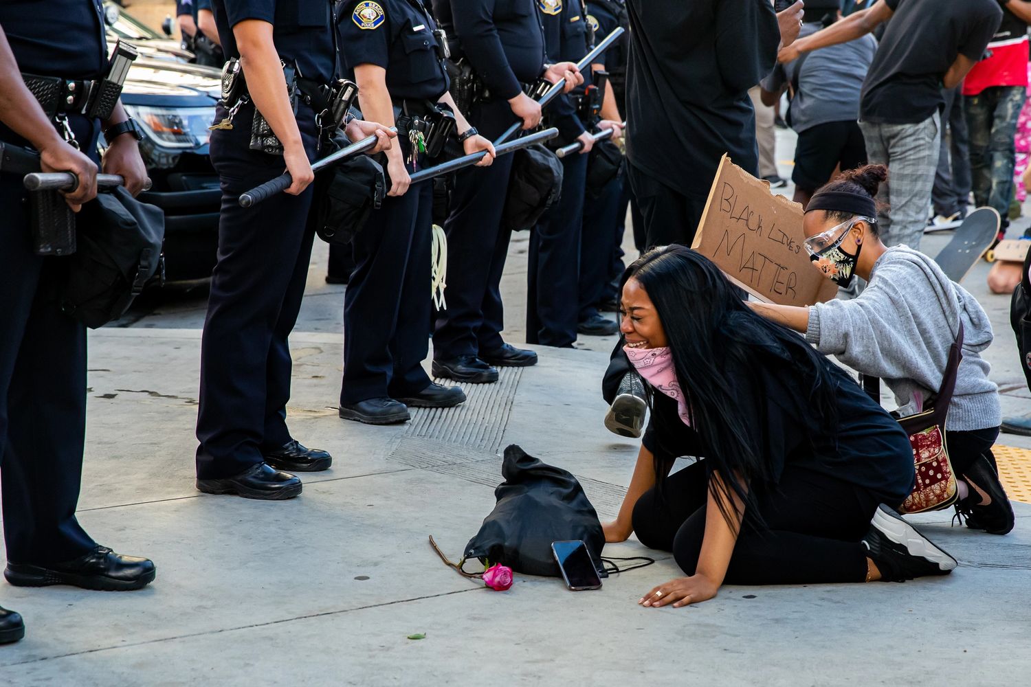A woman kneels in distress near police officers during a protest holding a Black Lives Matter sign.