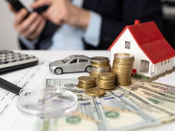 Stacks of coins, house model, and car on dollar bills representing financial planning.
