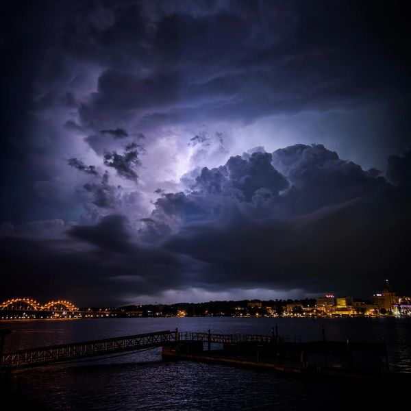 Dramatic lightning illuminates dark storm clouds over a city waterfront at night.