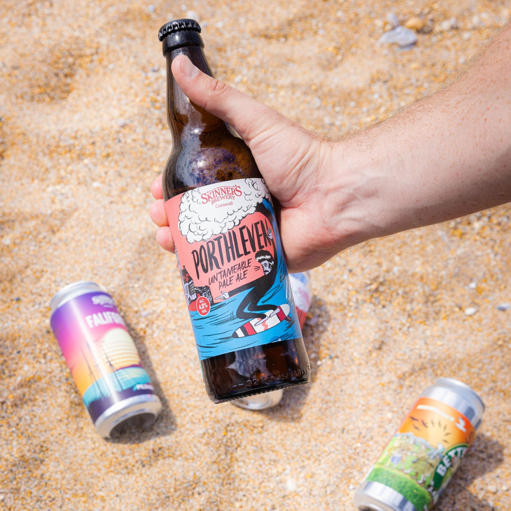 Hand holding a Porthleven Untameable Pale Ale bottle on a sandy beach.