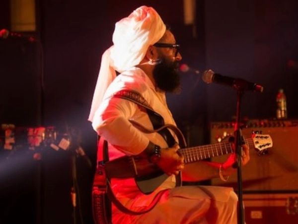 Bearded musician in traditional attire playing guitar on stage under warm lighting.
