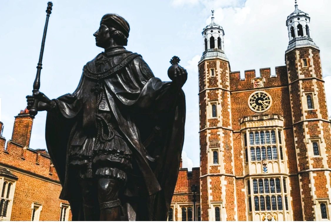 Statue of a regal figure with a scepter and orb in front of a historic brick building with towers.