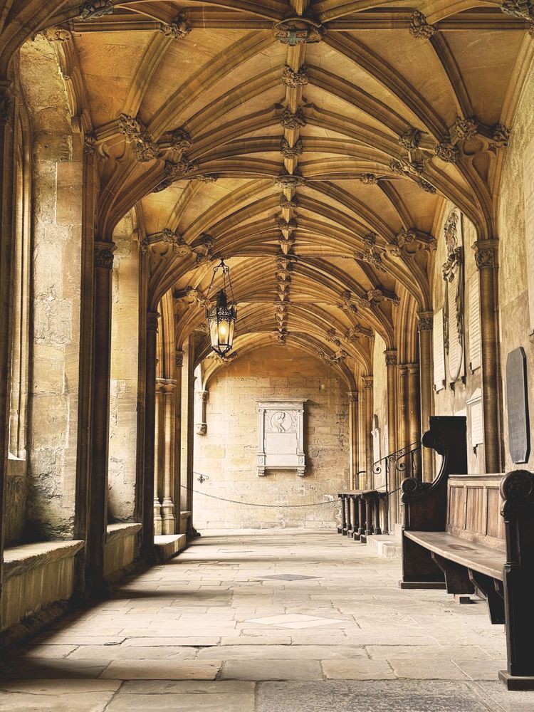 Ornate vaulted ceiling in a historic stone corridor with wooden benches and lantern.