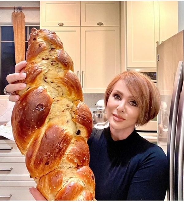 Woman proudly holding a large freshly baked braided bread in a kitchen.