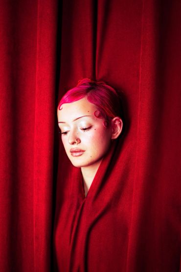 Woman with pink hair amidst red curtains.