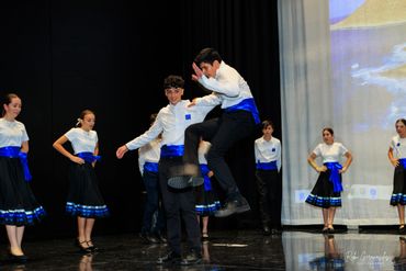 Young dancers perform a traditional group dance in coordinated blue and white costumes on stage.