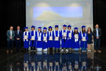 Graduates in blue gowns and caps holding certificates, standing with officials on stage.