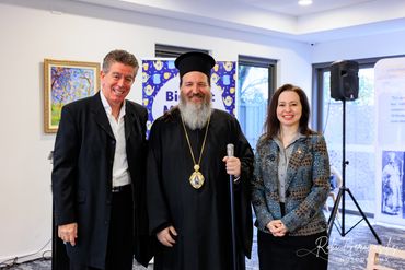 Three smiling adults posing indoors, including a religious figure in black robes with a staff.