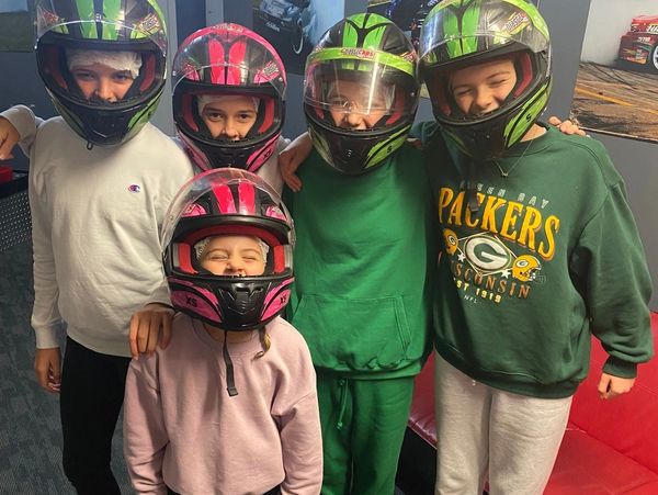 Four children wearing helmets and casual clothes pose together indoors.