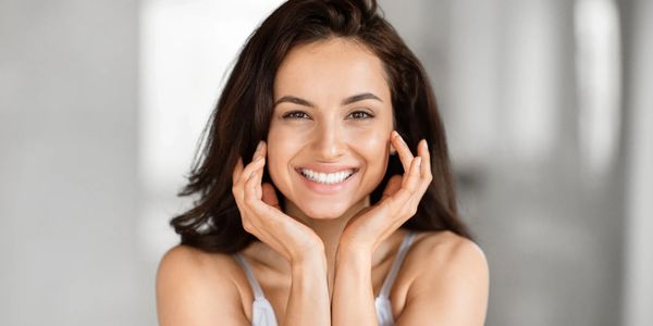 Young woman with glowing skin smiling brightly, hands framing her face.
