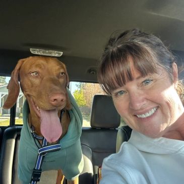 Woman and her dog smiling inside a car on a sunny day.