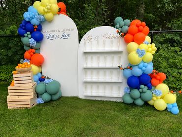 Colorful balloon arch surrounds a celebration setup with crates of oranges outdoors.