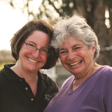 Woman with brown hair,  a black shirt and glasses smiling with a woman in a purple sweater with gray