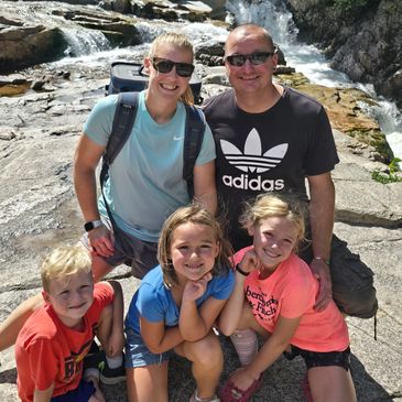 A smiling family of five enjoying a sunny day by a rocky river waterfall.