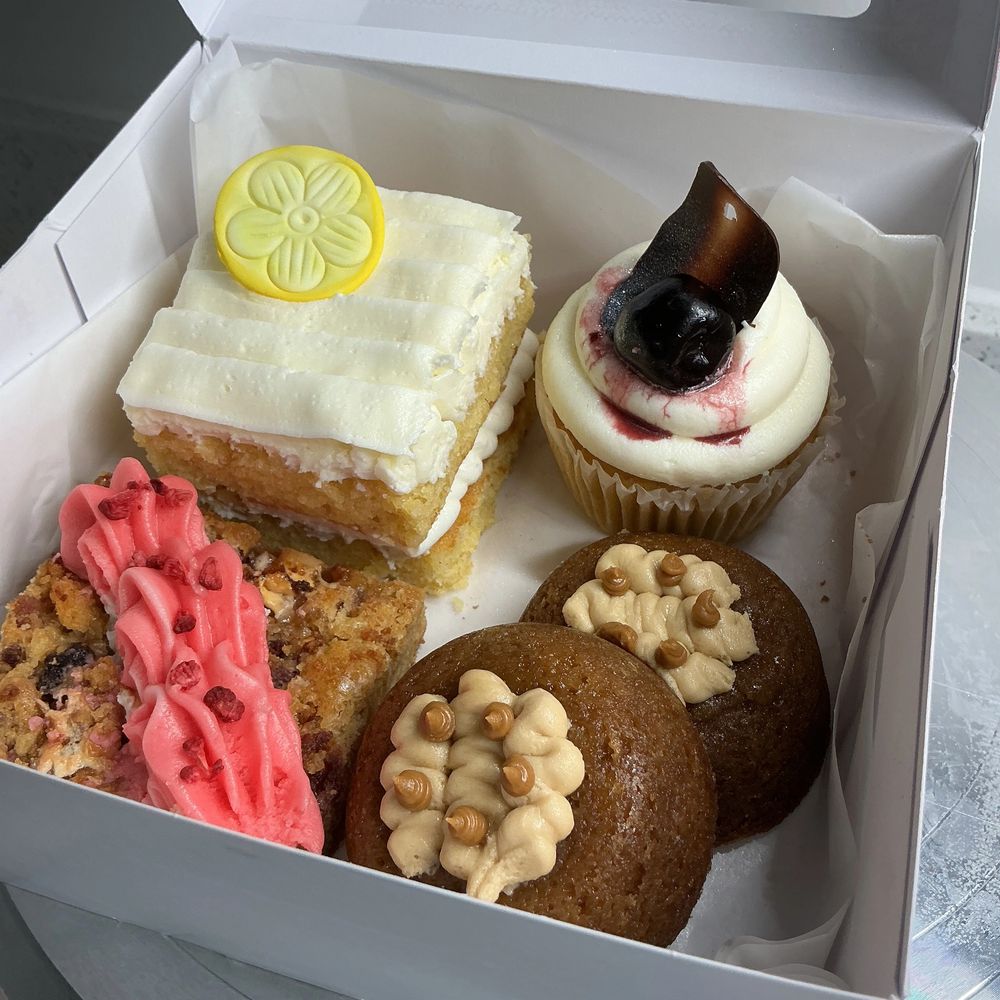 Assorted gourmet desserts in a white box, including cupcakes and bars with decorative frosting.
