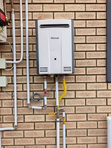 A Rinnai water heater mounted on a brick wall with connected pipes.