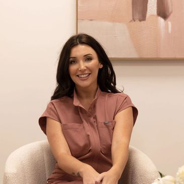 Smiling woman in a pink outfit sitting on a white chair indoors.